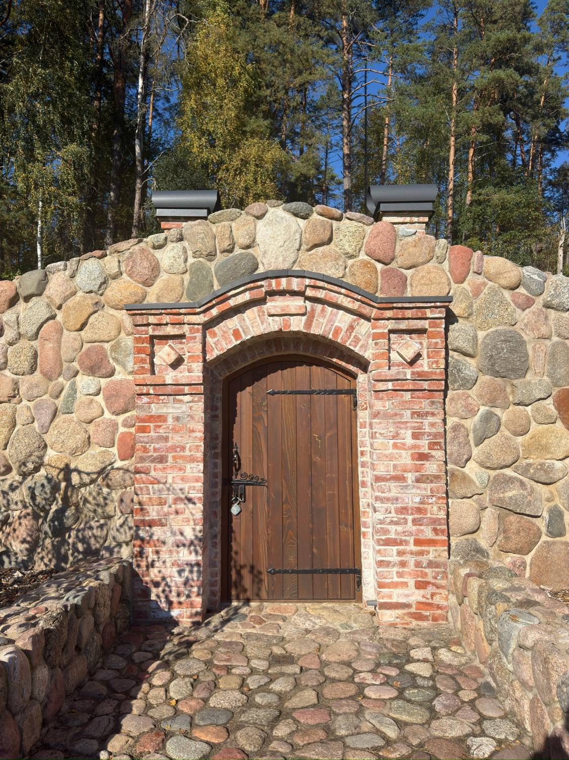Portal i återbrukat rött tegel med välvd båge och trädörr med smidda gångjärn, infälld i en mur av natursten, med kullersten framför